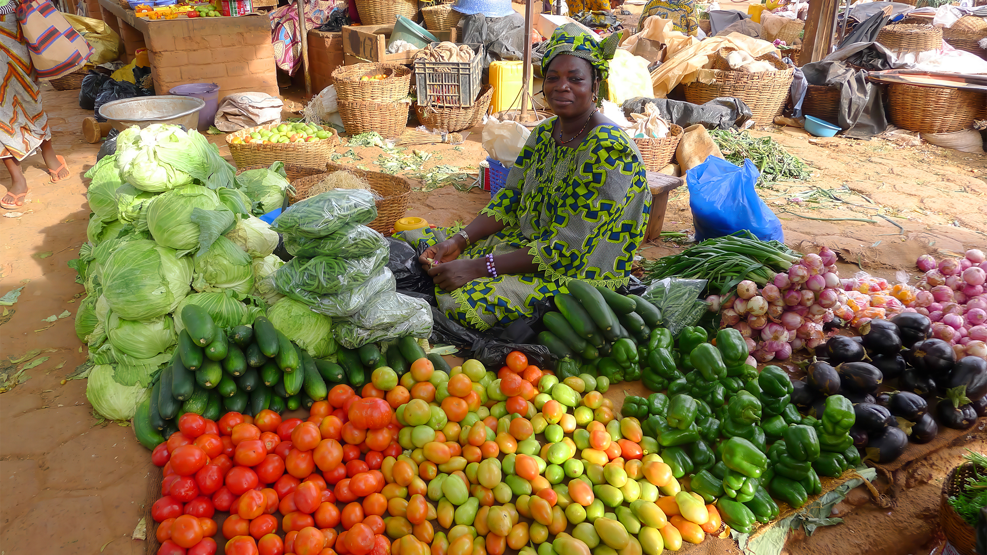 Mulher sentada num mercado tradicional rodeada por legumes frescos como alface, tomate, beringela, cebola e pimentos